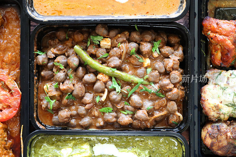 精选的印度外卖菜装在黑色塑料，一次性食品容器中，食谱包括，Pindi chole(鹰嘴豆咖喱)，Dal makhani, Palak奶酪，Chicken Bhuna, Murgh malai tikka(烤鸡)，高架视图